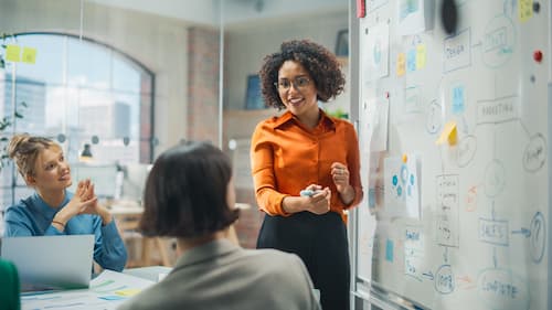 Woman standing next to a whiteboard conversing with team